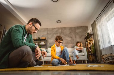 Anne kanepede otururken, baba ve oğul yerde domino oynuyorlar.