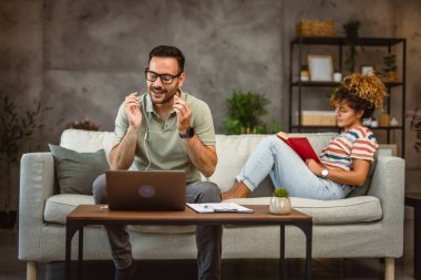 Kadın kanepede kitap okurken, erkek evde çalışırken müzik dinler.