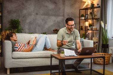 Kadın kanepede kitap okurken, erkek evde çalışırken müzik dinler.