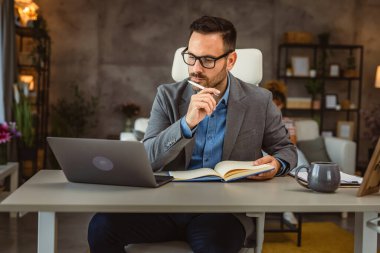 Yetişkin bir adam evde çalışıyor. Bilgisayardan kadın ve eşinin olduğu bir not defterine veri yazıyor.