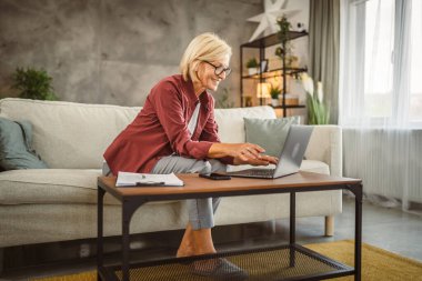 Evde dizüstü bilgisayarla çalışan olgun yaşlı bir kadın.