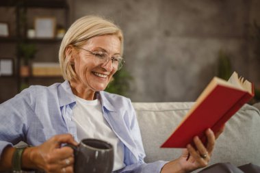 Olgun bir kadın oturma odasında oturur kitap okur ve kahve içer.
