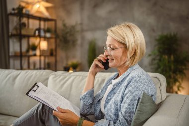 Olgun yaşlı kadın evde çalışıyor ve cep telefonuyla konuşuyor.