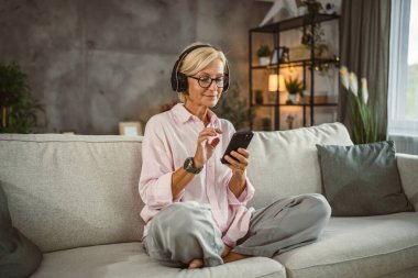 Olgun bir kadın, kulaklıkla koltuğa oturur ve cep telefonuyla müzik dinler.