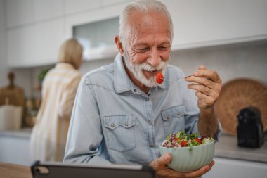 Yaşlı adam salata yiyip tablet izlerken olgun kadınlar kahvaltı hazırlıyor.