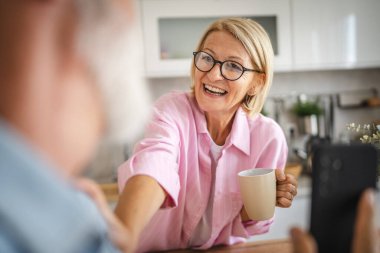 Omuz manzarası üzerinden yaşlı çift kocası mutfakta olgun karısına cep telefonu ekranını gösteriyor.