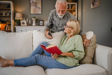 Olgun bir eş kanepeye uzanıp kitap okuyor. Kocasının yanında cep telefonuyla duruyor.