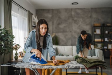 Yetişkin kadın, erkek tozları temizlerken gardırobu demir tahtada ütülüyor.