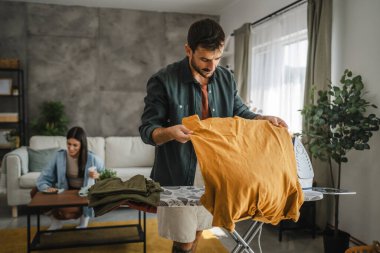 Yetişkin adam, kadın tozları temizlerken gardırobu demir tahtada ütülüyor.