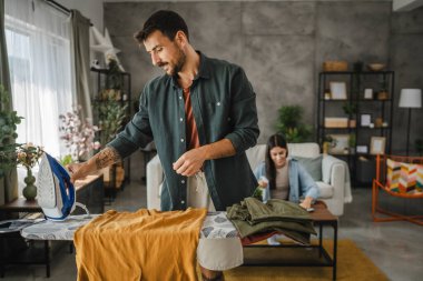 Yetişkin adam, kadın tozları temizlerken gardırobu demir tahtada ütülüyor.