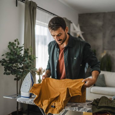 Yetişkin adam evdeki demir tahtanın üstündeki dolabı ütülüyor.