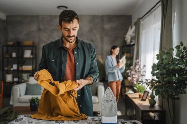 Yetişkin adam, kadın tozları temizlerken gardırobu demir tahtada ütülüyor.