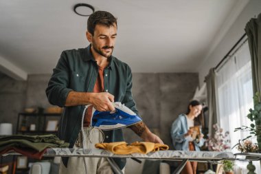 Yetişkin adam, kadın tozları temizlerken gardırobu demir tahtada ütülüyor.