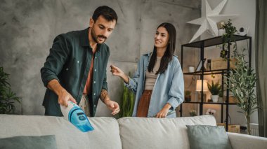 Yetişkin bir adam elektrikli süpürgeyle kanepeyi süpürürken bir kadın onu destekliyor.