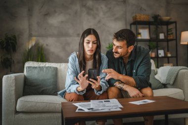 Adult young woman prepare to have contactless payment scan invoice on cellphone while her boyfriend sit next to her on sofa clipart