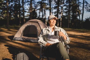 Genç bayan çadırın önünde oturur ve ormanda bir fincan kahve ya da sıcak bir şeyler içer.