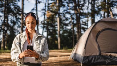 Kulaklıklı genç bir kadın yere oturup cep telefonu kullanıyor ve dağda müzik dinliyor.