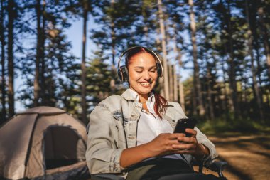 Kulaklıklı genç bir kadın çadırın önünde oturur ve cep telefonu kullanır, dağda müzik dinler.