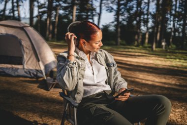 Kulaklıklı genç bir kadın çadırın önünde oturur ve cep telefonu kullanır, dağda müzik dinler.