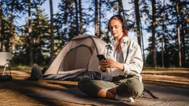 Kulaklıklı genç bir kadın yere oturup cep telefonu kullanıyor ve dağda müzik dinliyor.