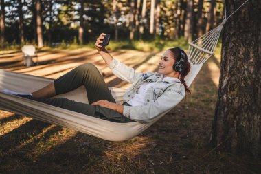Hamakta kulaklıkla uzanan genç bir kadın cep telefonunu kendi portresi ya da video görüşmesi için kullanıyor.