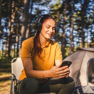 Kulaklıklı genç bir kadın çadırın önünde oturur ve cep telefonu kullanır, dağda müzik dinler.
