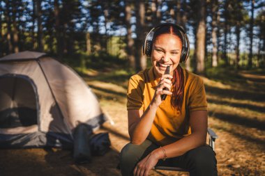 Kulaklıklı genç bir kadın önde oturur müzik dinler ve cep telefonuyla şarkı söyler.