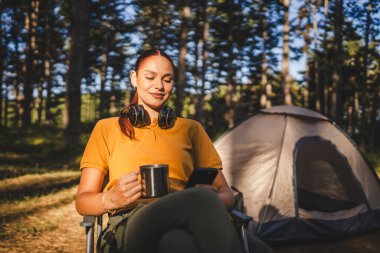 Genç bayan çadırın önünde oturur, cep telefonu kullanır ve kahve içer.