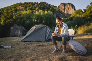 Genç bayan çadırın önünde oturur ve ormanda cep telefonu kullanır.