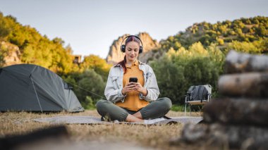 Genç kadın bacak bacak üstüne atarak oturur ve kulaklıkla müzik dinler ve ormanda cep telefonu kullanır.