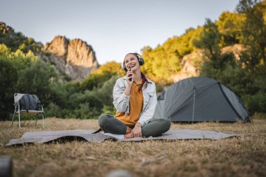 Genç kadın bacak bacak üstüne atmış bir şekilde battaniyeye oturur kulaklıklarla müzik dinler ve cep telefonuyla şarkı söylerdi.