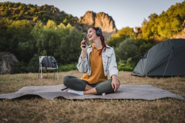 Genç kadın bacak bacak üstüne atmış bir şekilde battaniyeye oturur kulaklıklarla müzik dinler ve cep telefonuyla şarkı söylerdi.