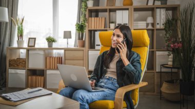 Yetişkin kadın evde çalışıyor cep telefonuyla konuşuyor ve dizüstü bilgisayar kullanıyor.