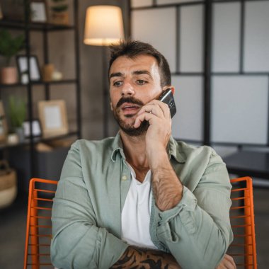 Öfkeli bir yetişkinin portresi sandalyeye oturup cep telefonuyla konuşur.