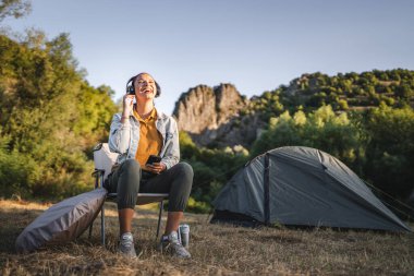 Genç bayan kulaklıkla müzik dinler, çadırın önüne oturur, ormanda cep telefonu kullanır.