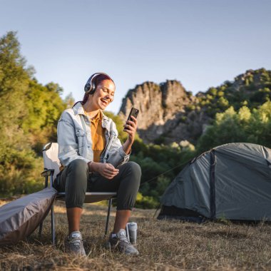 Genç bayan kulaklıkla müzik dinler, çadırın önüne oturur, ormanda cep telefonu kullanır.