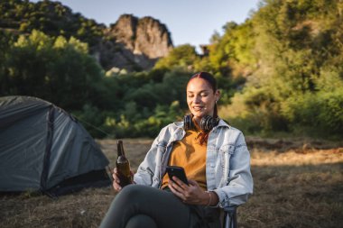 Kulaklıklı genç bir kadın çadırın önünde oturur ve bira içer. Ormanda cep telefonu kullanır.