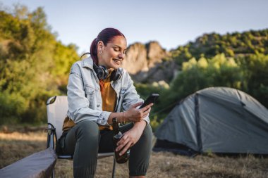 Kulaklıklı genç bir kadın çadırın önünde oturur ve bira içer. Ormanda cep telefonu kullanır.