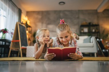 İki kız arkadaş, halıya uzanmış kız kardeş ve bir kitap okuyorlar.