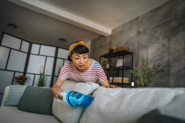 Olgun Japon kadın elektrikli süpürgeyle kanepeyi süpürüyor ev işleri yapıyor.