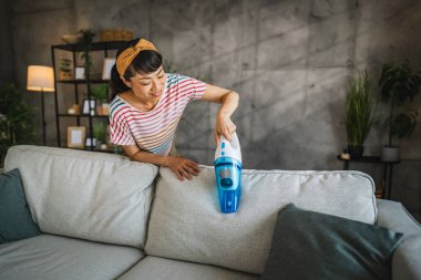 Olgun Japon kadın elektrikli süpürgeyle kanepeyi süpürüyor ev işleri yapıyor.