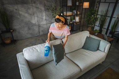 Olgun Japon kadın elektrikli süpürgeyle kanepeyi süpürüyor ev işleri yapıyor.