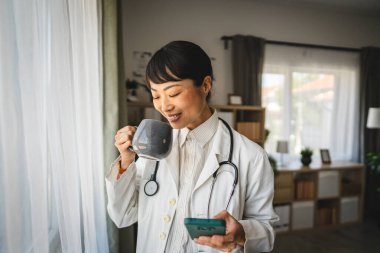 Olgun Japon kadın doktor iş yerindeki cep telefonuyla kahve molası verdi.