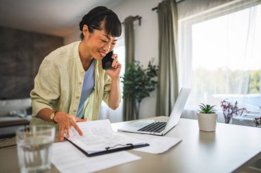 Ev bilgisayarında çalışan ve cep telefonuyla konuşan Japon bir kadın.