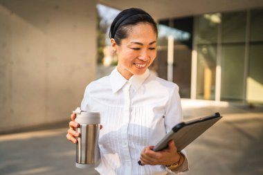 Olgun Japon iş kadını dijital tablet kullanıyor ve termosu modern binaların önünde tutuyor.