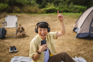 Neşeli, kulaklıklı, olgun Japon kadın kamp gezisinde çadırın önünde oturur ve cep telefonuyla müzik dinler.