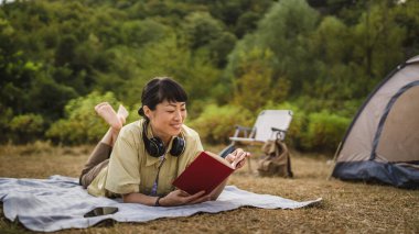 Olgun Japon kadın yere uzanıp kamp gezisi üzerine bir kitap okuyor.