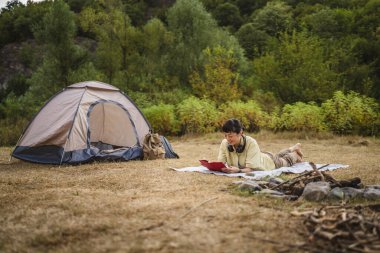 Mature japanese woman lying down on a ground and read a book on camping trip clipart