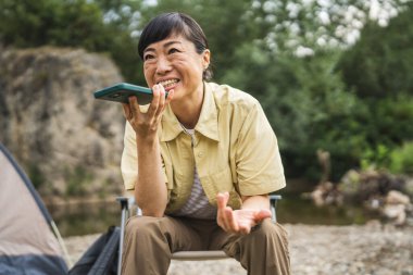 Mature japanese woman sit in front of tent and record voice mail, audio message on mobile phone clipart