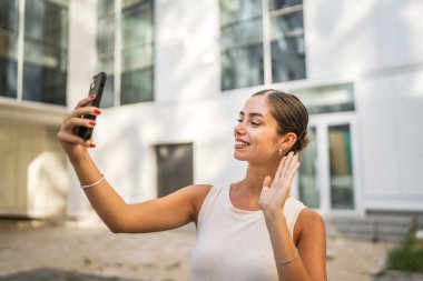 Genç bir kadın binanın önünde duruyor ve cep telefonuyla video görüşmesi yapıyor.